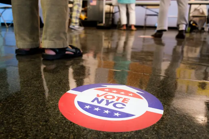 A NYC Votes sticker on the floor, with people's legs and feet in the background
